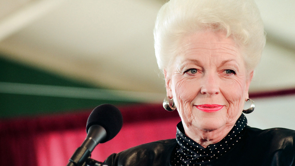 Ann Richards stands, smiling, behind a microphone