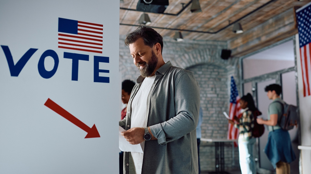 A smiling man stands next to a sign with an American flag and the word Vote. He is about to cast his ballot, as others stand behind him waiting for their turn to vote.