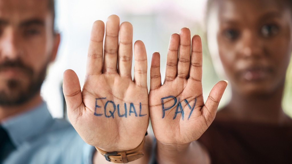 A man and woman each holds a palm up to face the camera. The man is on the left with the word “equal” written on his hand, and the woman is on the right with the word “pay” written on hers