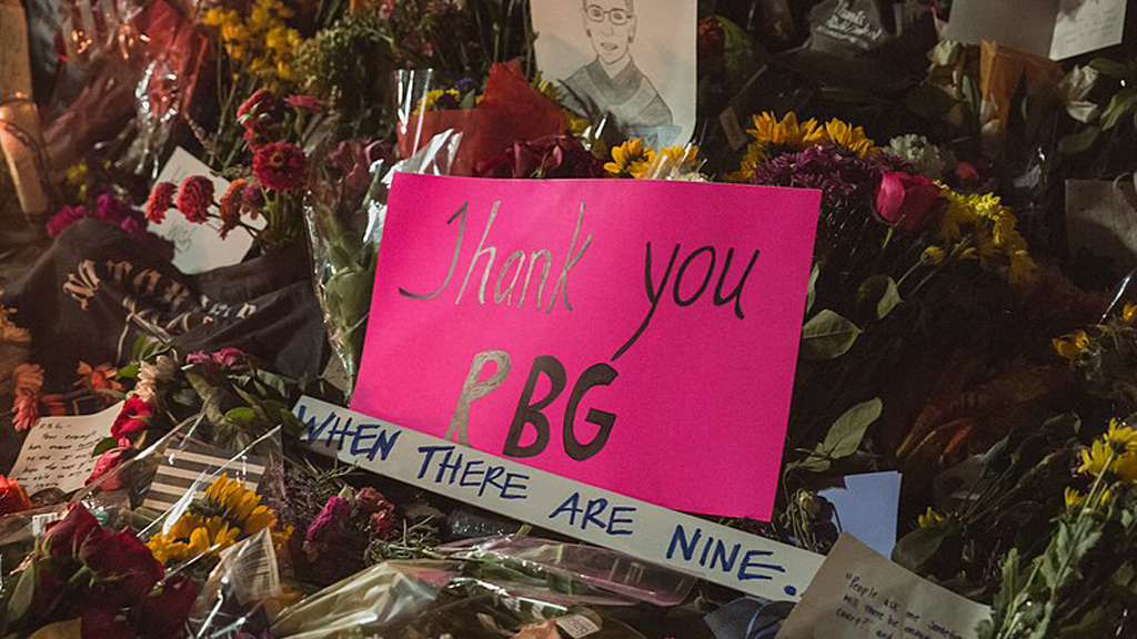 A vibrant pink sign reading 'Thank you RBG' is surrounded by flowers and tributes. A hand-drawn picture and messages are also visible in the background