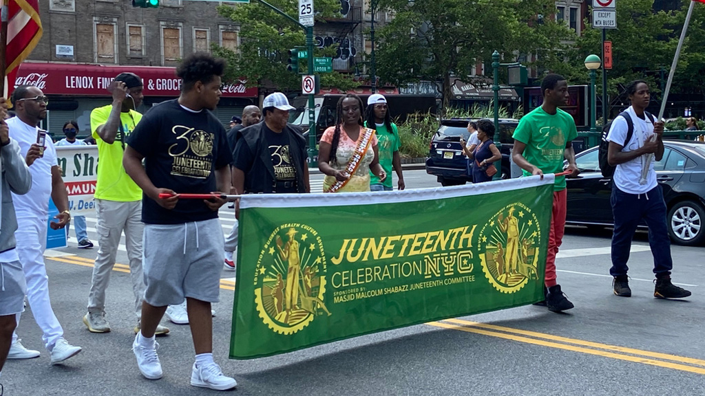 A group of people in a Juneteenth parade carry a green banner with yellow text that says, Juneteenth Celebration NYC. They walk along a street named Malcolm X Boulevard, passing storefronts and a traffic light