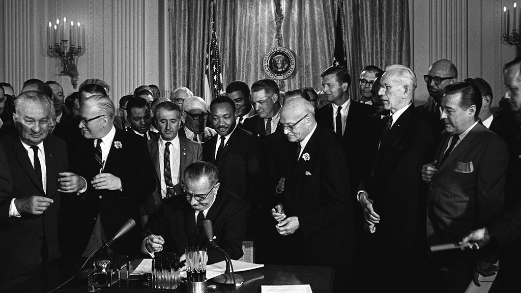 President Johnson sits behind a desk at the White House. There are many men crowded behind him, as he signs the Civil Rights Act into law. Many pens are lined up in front of him, soon to be handed out as keepsakes