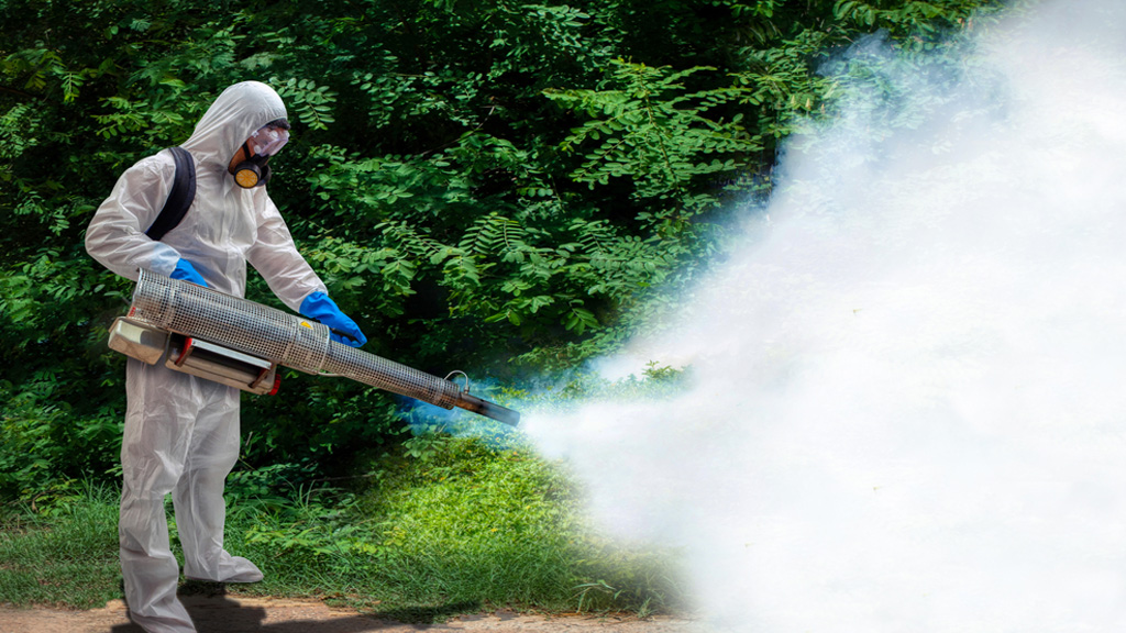 A man dressed completely in protective gear holds a large canister that he uses to spray a large cloud of pesticide towards a wooded area