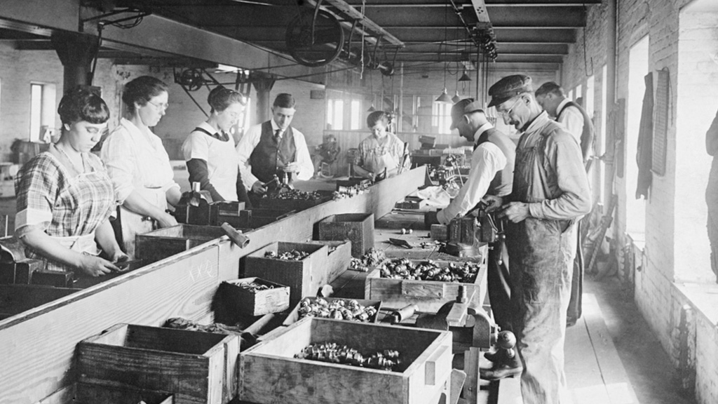 A black and white photograph shows men and women working on either side of an assembly line in 1920. They are making spark plugs.