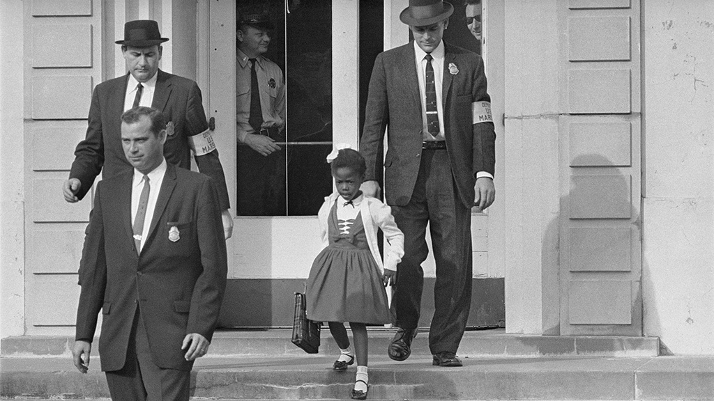 A small black girl walks down the steps of her school surrounded by five white men to protect her