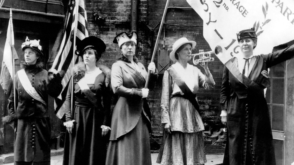 Five women in early 20th century attire hold suffrage banners and flags