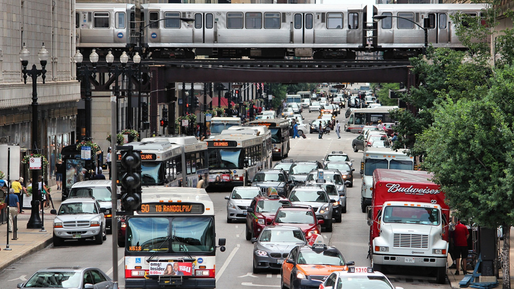 A busy street scene shows pedestrians, buses, cars, taxis, delivery trucks, and an elevated train.