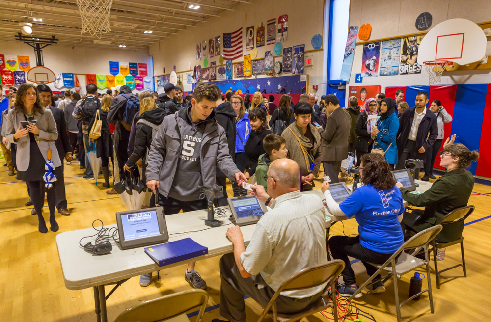 A large crowd lines up to vote inside a school gym. Three election officials sit at a table, checking photo IDs.